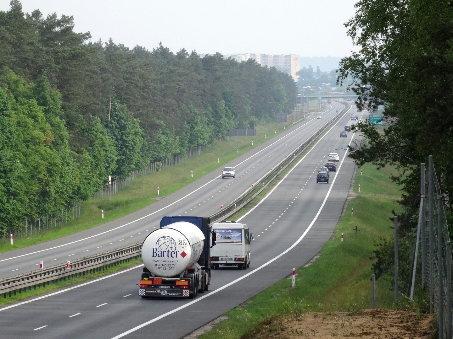 Ein großer Lkw fährt auf einer von beiden Seiten mit Bäumen gesäumten Autobahn, mit einem Zaun und Gras auf der rechten Seite und im Hintergrund sichtbare Gebäude unter einem klaren blauen Himmel.