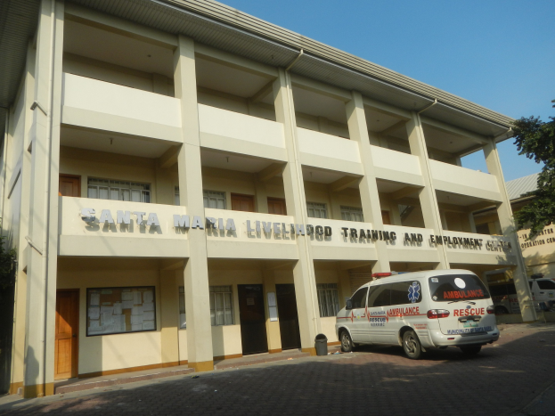 Lebensmittel-Trainings- und Arbeitsplatz-Zentrum Santa Maria mit Fenstern, Türen und Text umgeben von Bäumen und Fahrzeugen auf der Straße unter einem klaren blauen Himmel.