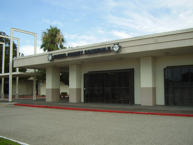 Außenansicht des Harris County Constable's Office-Gebäudes mit Glaswänden und Säulen, einer Straße, Gras, Bäumen und einem bewölkten Himmel im Hintergrund.