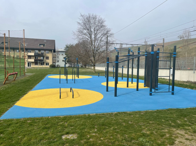 Spielplatz mit einem blauen und gelben Spielbereich, umgeben von Gras, Zäunen, Bäumen, Gebäuden, Versorgungsmasten und einem bewölktem Himmel.
