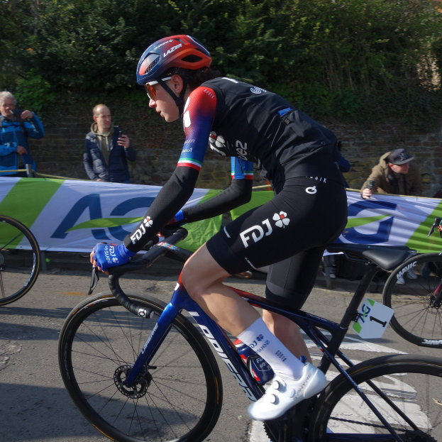Die professionelle Radrennfahrerin Katie Archibald fährt ein Fahrrad auf einer Straße, trägt einen Helm, eine Brille, Handschuhe und Schuhe und ist von einer Menschenmenge umgeben. Im Hintergrund gibt es ein Banner mit Text, eine Wand, Bäume und ein Gebäude.