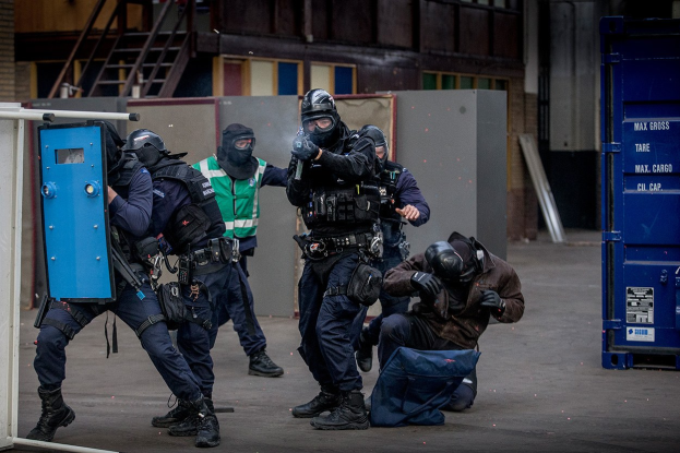 Polizeibeamte in Einsatzausrüstung stehen vor einem Gebäude, einige halten Waffen, mit einer Person am Boden im Vordergrund und einem blauen Objekt mit Text auf der rechten Seite.