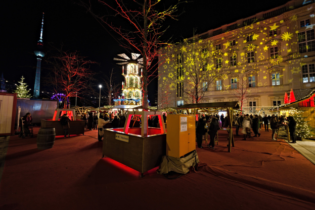 Ein belebter Weihnachtsmarkt in Berlin, Deutschland mit Menschen um geschmückte Stände, festliche Lichter, Bäume, Gebäude, Laternenmasten und einen Turm unter einem dunklen Himmel.