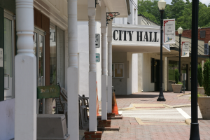 Ein Rathaus umgeben von Gebäuden, Straßenlaternen, Straßenschildern, Kraftfahrzeugen, Verkehrskegeln, Stühlen, Topfpflanzen, Bäumen und einem bewölkten Himmel.