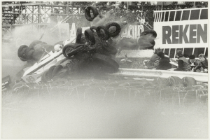 Ein Schwarz-Weiß-Foto eines verunglückten Rennwagens, umgeben von Reifen, mit einer Gruppe von Menschen rechts und einer Texttafel im Hintergrund.