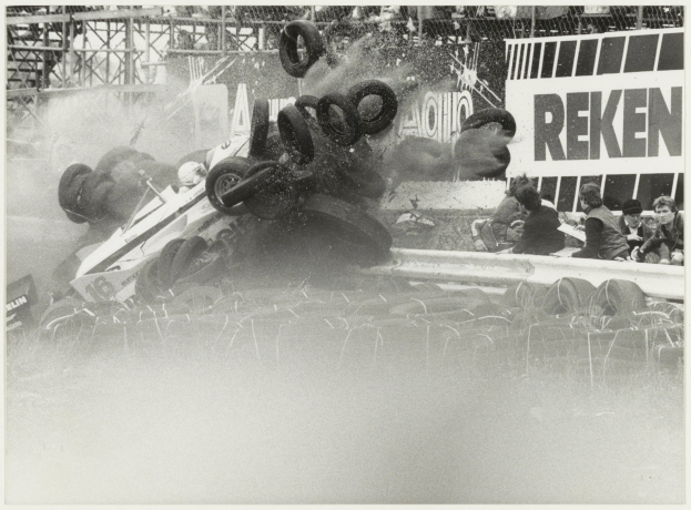 Ein Schwarz-Weiß-Foto eines verunglückten Rennwagens, umgeben von Reifen, mit einer Gruppe von Menschen rechts und einer Texttafel im Hintergrund.