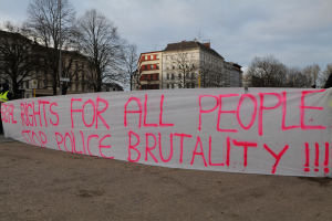 Eine Gruppe von Menschen, die auf dem Boden stehen und ein Transparent halten, auf dem 'Rechte für alle Menschen Stoppt Polizeigewalt' steht, mit einem Straßenschild, einem Schild, Bäumen, Gebäuden mit Fenstern und einem bewölkten Himmel im Hintergrund.