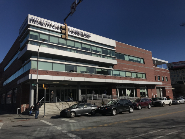 Großes Gebäude mit der Bezeichnung New York City Health Care Homeless Center mit geparkten Autos, Fußgängern auf einem Gehweg, Straßenmöblierung und Bäumen unter einem klaren Himmel.