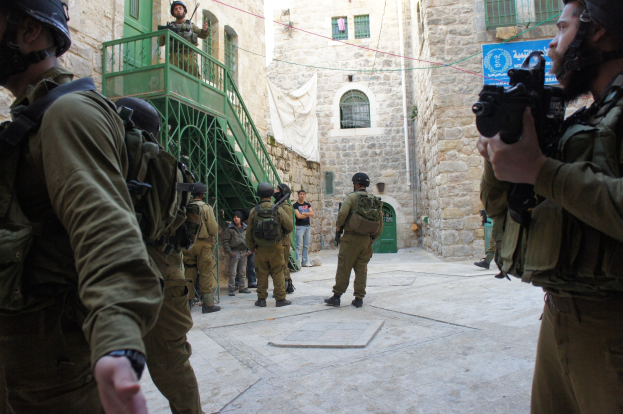 Israeli soldiers in helmets and gear stand in Jerusalem's old city, some holding rifles, with buildings and a banner in the background.
