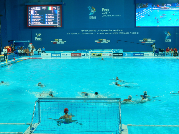 Eine Gruppe von Menschen, die Wasserball in einem großen Pool mit einem Netz in der Mitte spielen, Zuschauer auf der linken Seite und ein Bildschirm im Hintergrund mit Text und Logos bei den FINA-Weltmeisterschaften 2016 in Kazan.