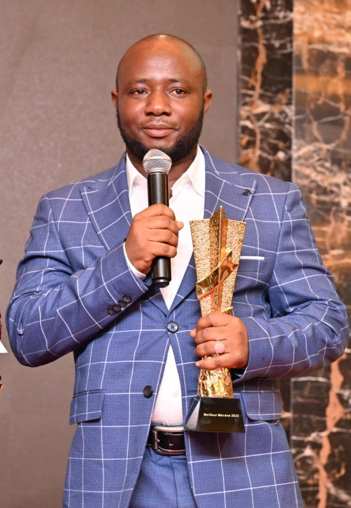 Ein Mann in einem blauen Anzug, der ein Mikrofon und eine Trophäe hält und mit einem glücklichen Ausdruck und selbstbewusster Haltung feiert.