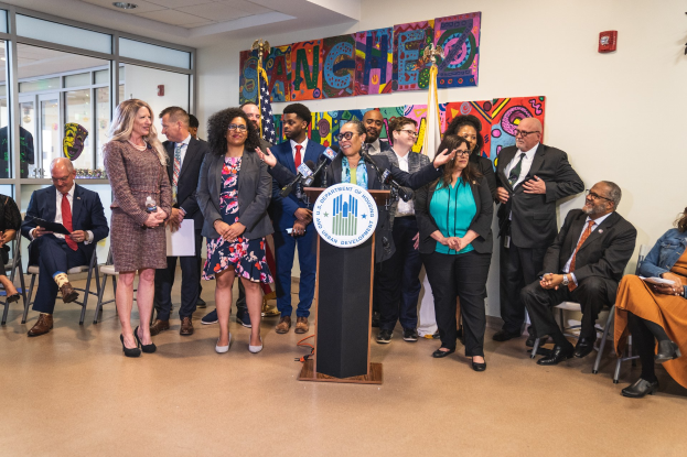 Gruppe von Menschen vor einem Podium mit einem Mikrofon und einer Tafel mit der Aufschrift "Sancho City Councilman Joins City Council", mit sitzenden und stehenden Zuschauern, Flaggen, einem Feuerlöscher und Deckenlampen im Hintergrund.