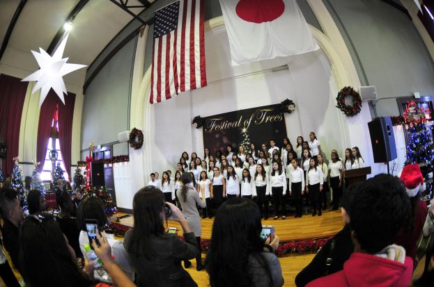 Gruppe von Menschen auf einer Bühne mit Weihnachtsdekorationen, einem Podium, Lautsprechern, einer Texttafel, Vorhängen, Flaggen und Deckenleuchten vor einem Publikum mit Kameras und Handys.