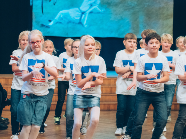 Eine Gruppe von Kindern in weißen Hemden und blauen Shorts, einige mit Brille, tanzt auf einer Bühne während eines Wettbewerbs, mit Zuschauern auf der linken Seite und einem Bildschirm im Hintergrund.