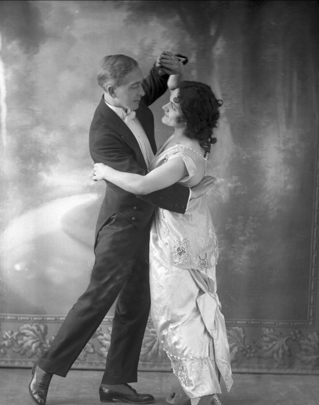 Schwarzes und weißes Foto eines Mannes in einem Anzug und einer Frau, die Tango tanzen, mit kunstvollen Wanddesigns im Hintergrund.