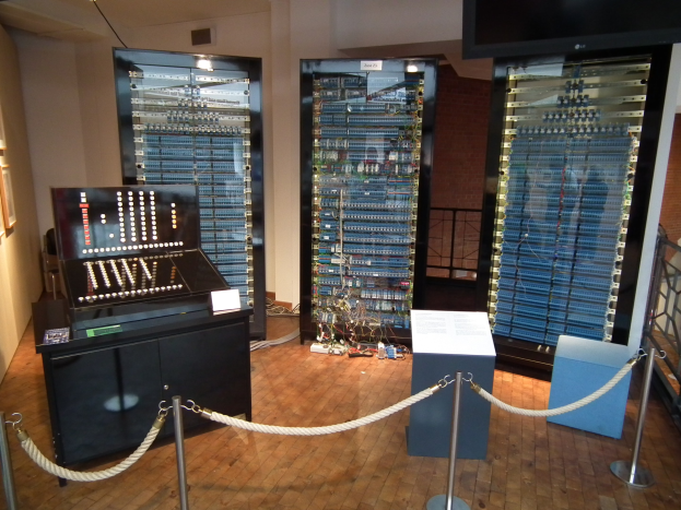 Ein unordentlicher Raum mit verschiedenen elektronischen Geräten, ein Tisch mit Gegenständen, eine bodengeständiges Texttafel, Absperrbänder an Pfählen, ein Metallzaun, gerahmte Bilder an der Wand und Deckenleuchten.