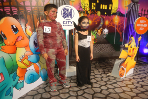 Zwei Kinder in Halloween-Kostümen stehen nebeneinander vor einer Wand mit einem Bild eines Gebäudes, Bäumen, Laternenmasten und einem Zaun sowie Schildern mit Text und Tierbildern und festlicher Beleuchtung.