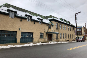 Ein großes Backsteingebäude mit einem grünen Dach an einer Straßenecke, identifiziert als die ehemalige Feuerwache St. Louis, umgeben von Fahrzeugen, Schnee, Strommasten, Bäumen und einem bewölkten Himmel.
