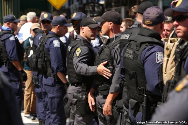 Gruppe von Polizisten in Uniform auf einer Straße stehend, einige halten Waffen, mit Gebäuden und Texttafeln im Hintergrund.