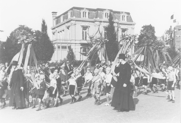 Schwarzes und weißes Bild einer Gruppe von Menschen, die eine Straße entlanggehen, einige halten Fahnen, mit Hüten auf dem Kopf, vorbei an einem Gebäude und Bäumen, unter einem sichtbaren Himmel, während eines Maifeiertags mit einem Maibaum.