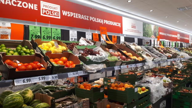 Ein Ladeneinrichtungsbild, das verschiedene Obst- und Gemüsesorten in Kartonschachteln mit Preisschildern zeigt, mit informativen Tafeln an den Wänden und Deckenbeleuchtung.