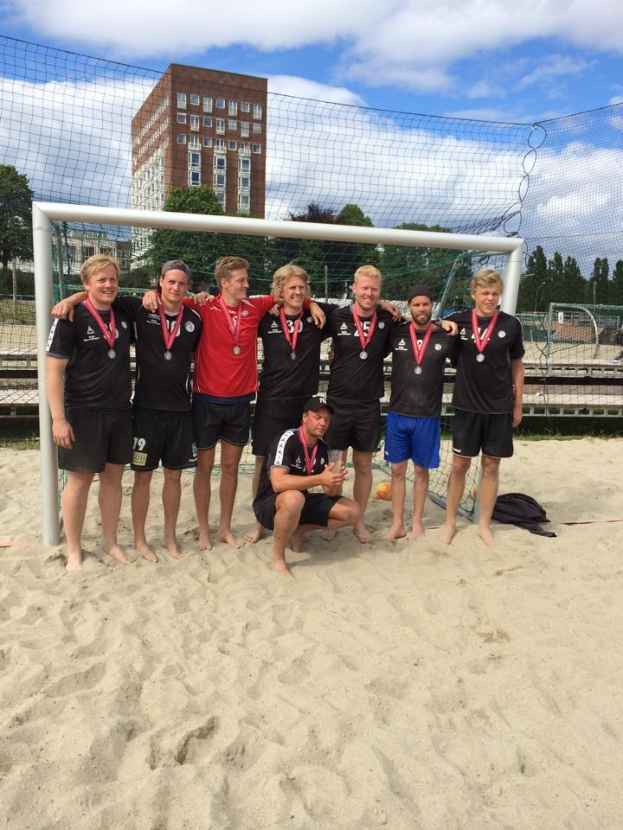 Mannergruppe in Sportkleidung auf einem sandigen Strand, mit Medaillen ausgestattet und von einem Netz umgeben, mit Bäumen, Gebäuden und einem bewölkten Himmel im Hintergrund.