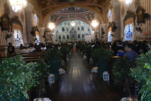 Eine Hochzeitszeremonie in einer Kirche mit Sitzenden und Stehenden, Topfpflanzen im Gang, verzierten Wänden, Vorhängen an den Fenstern, Kronleuchtern und einem Bildschirm im Hintergrund.