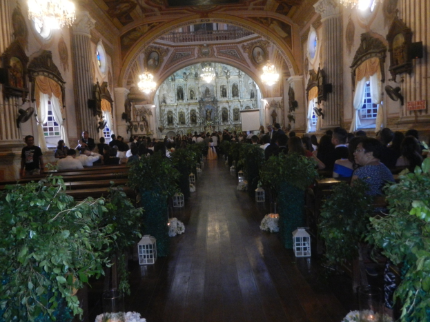 Eine Hochzeitszeremonie in einer Kirche mit Sitzenden und Stehenden, Topfpflanzen im Gang, verzierten Wänden, Vorhängen an den Fenstern, Kronleuchtern und einem Bildschirm im Hintergrund.