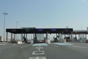 Mautstelle an der Straße mit einem vor ihr geparkten Lastwagen, weiße und blaue Straßenmarkierungen, Laternenpfähle, Schilder, Gebäude, Bäume und einen klaren blauen Himmel im Hintergrund.