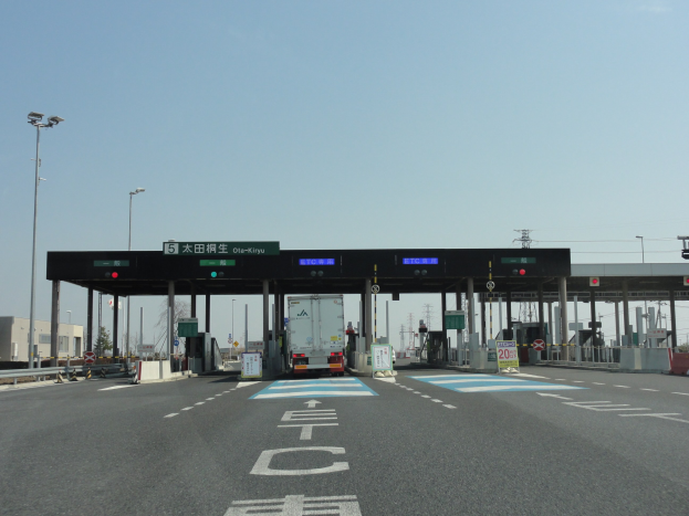 Mautstelle an der Straße mit einem vor ihr geparkten Lastwagen, weiße und blaue Straßenmarkierungen, Laternenpfähle, Schilder, Gebäude, Bäume und einen klaren blauen Himmel im Hintergrund.