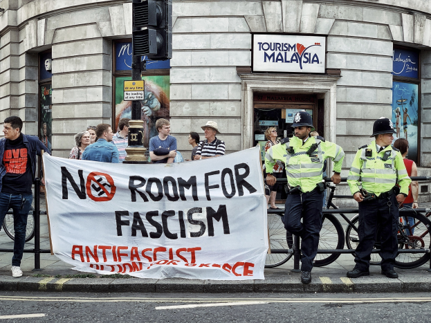 Gruppe von Menschen auf einem Fußweg mit einer 'Kein Platz für Faschismus'-Plakette, zwei Polizeibeamte vorne, Fahrräder, eine Ampel und ein Gebäude im Hintergrund.