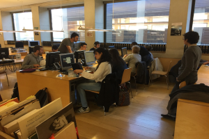 Eine Gruppe von Menschen, die an Computern auf Schreibtischen in einer Bibliothek arbeiten, umgeben von Bücherregalen, Monitoren, Tastaturen, Mäusen, Taschen auf dem Boden, Deckenlampen und Glasfenstern im Hintergrund.