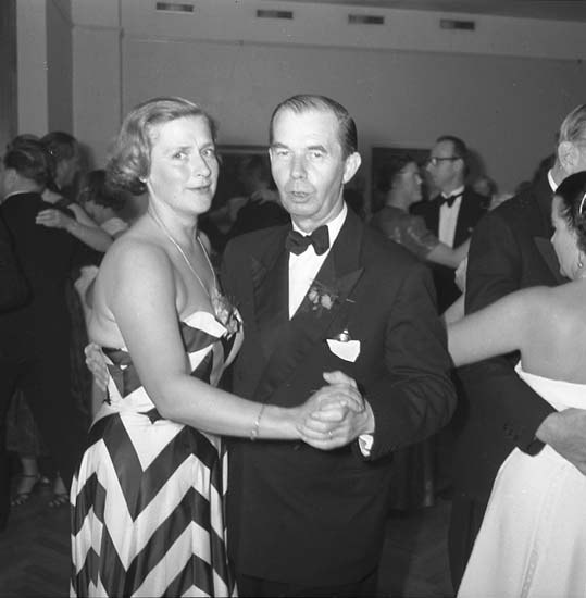 Ein Mann in einem schwarzen Blazer, weißem Hemd und schwarzen Hosen tanzt mit einer Frau in einem unbekannten Outfit auf einer formellen Veranstaltung, indem sie sich an den Händen halten und zur Musik schwenken, vor einer Wand als Hintergrund.