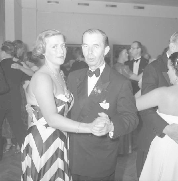 Ein Mann in einem schwarzen Blazer, weißem Hemd und schwarzen Hosen tanzt mit einer Frau in einem unbekannten Outfit auf einer formellen Veranstaltung, indem sie sich an den Händen halten und zur Musik schwenken, vor einer Wand als Hintergrund.