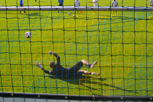 Ein Fußballspieler macht einen gehaltenen Ball vor einem Tor, mit einem Netz im Vordergrund und einigen Menschen im Hintergrund, sowie mehreren Bällen auf dem Boden verteilt.