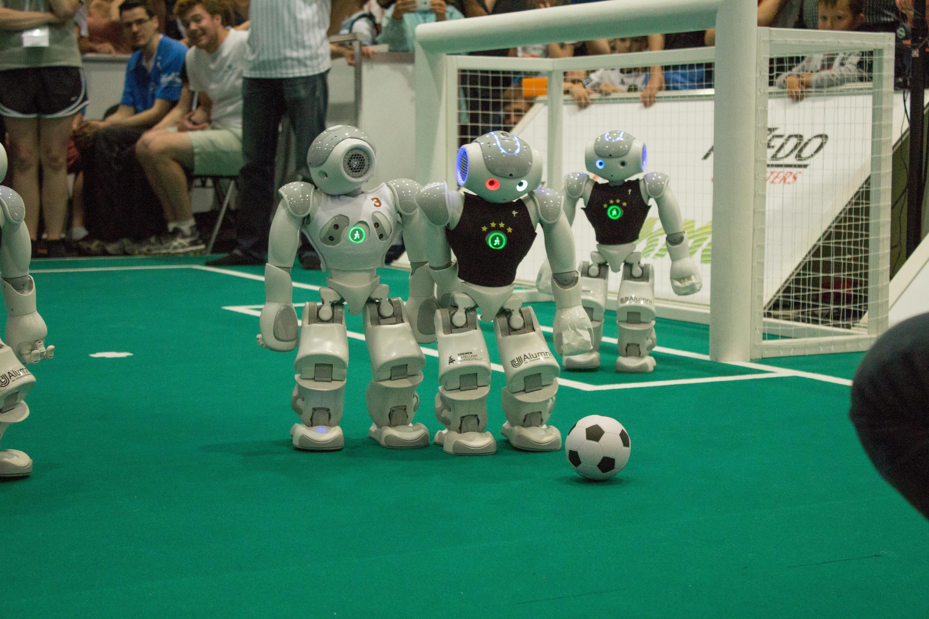 Eine Gruppe von Robotern, die auf einem grünen Feld Fußball spielen, mit einem Ball in der Mitte und einem Netz auf der rechten Seite sowie Zuschauern mit Kameras im Hintergrund.