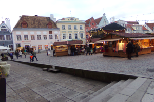 Ein lebendiger Weihnachtsmarkt in Tallinn, Estland, mit Menschen um beleuchtete und dekorierte Stände, Gebäuden mit Fenstern im Hintergrund, Treppen, die zum Markt führen, und Töpfen mit Pflanzen, die herumstehen.