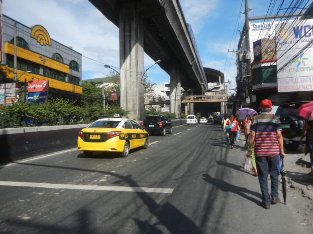 Ein gelbes Taxi fährt auf einer Straße mit hohen Gebäuden, Fußgängern mit Regenschirmen, einer Brücke im Hintergrund, bewölktem Himmel und Stromleitungen oben.