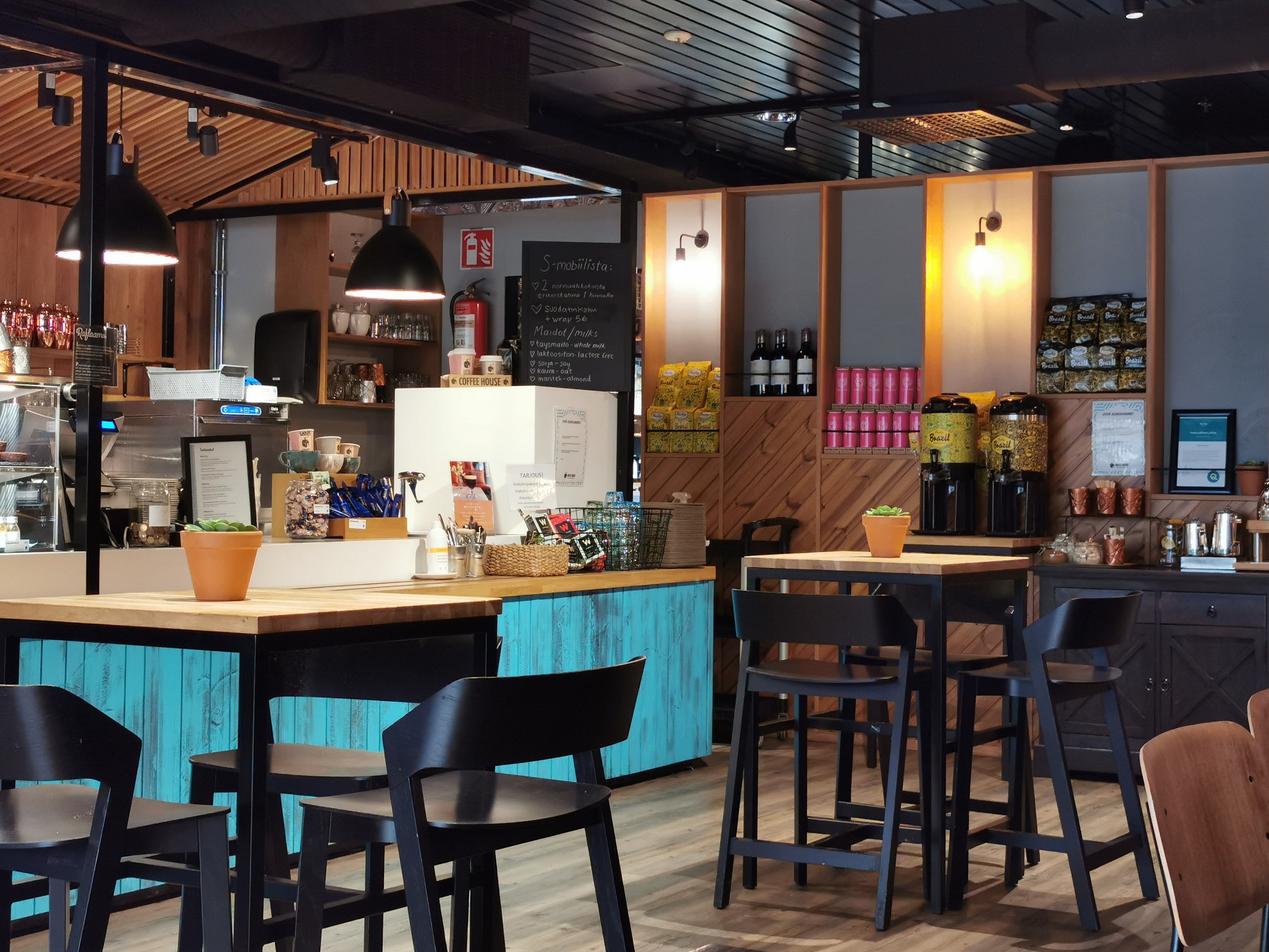 Innenraum eines Restaurants mit Holztischen, -stühlen, einer Topfpflanze auf einem Tisch, einer Schiefertafel, einem Kühlschrank, einem Feuerlöscher, Regalen mit Flaschen, Schränken mit Gegenständen, einem Rahmenfoto an der Wand und Deckenleuchten.