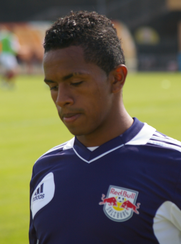 Ein junger Mann in einem blauen T-Shirt steht auf einem Fußballfeld und sieht aus wie ein Flügelspieler mit kurzen, dunklen Haaren und einem entschlossenen Gesichtsausdruck.