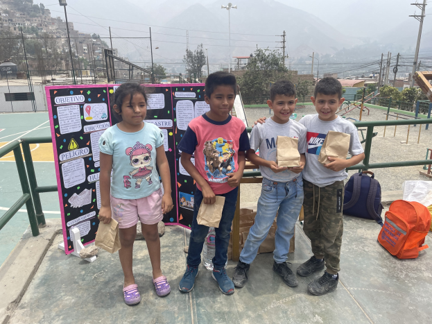 Gruppe von Kindern auf einem Basketballfeld mit Papiertüten in den Händen, mit Vorräten auf dem Boden, einer Tafel mit Text links und einem Hintergrund aus Zaun, Pfosten, Straßenlaternen, Bäumen, Gebäuden, Hügeln und Himmel.