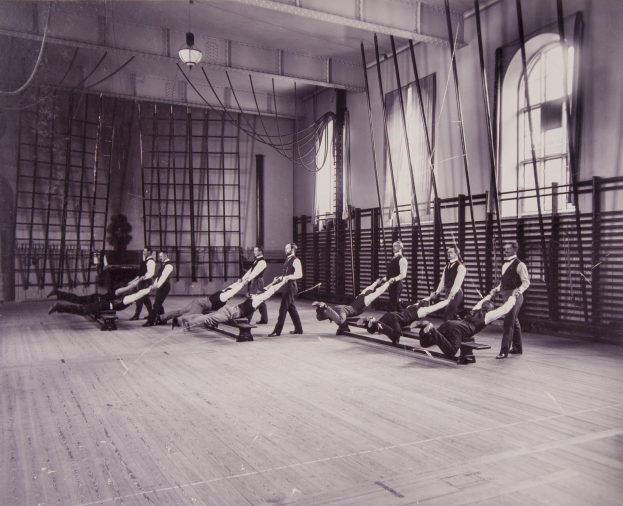 Schwarzes und weißes Bild einer Gruppe von Menschen, die Pilates-Übungen in einem Fitnessstudio mit einer Wand, Fenstern auf der rechten Seite und Deckenlampen im Hintergrund machen.