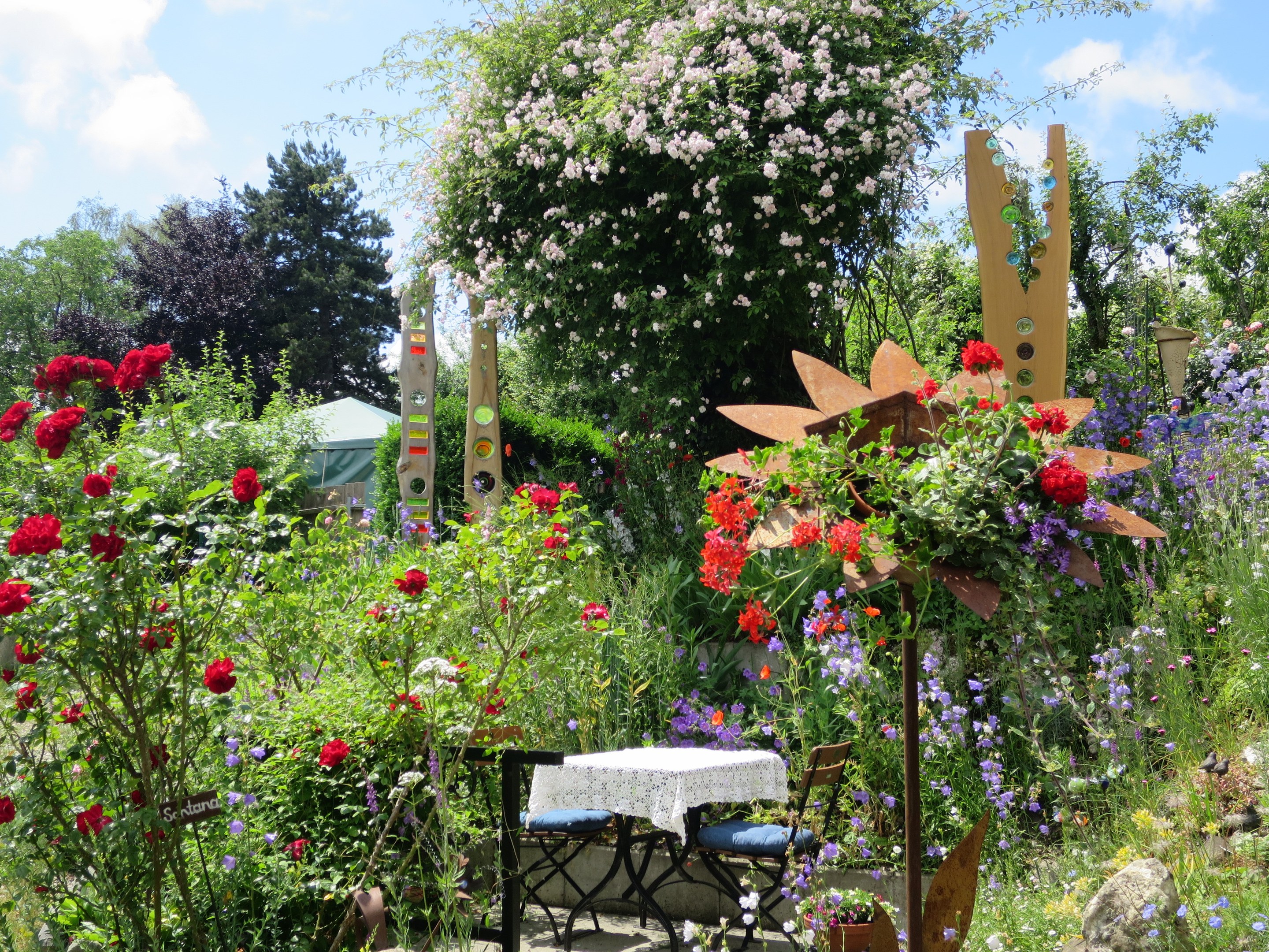 Ein Garten mit bunten Blumen und Pflanzen, der einen Tisch mit einer Decke und Stühlen in der Mitte und umgeben von Bäumen unter einem bewölkten Himmel auf dem Chelsea Flower Show zeigt.