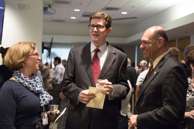 Eine Gruppe von Menschen in einem Raum, mit einem Mann in einem Anzug, der ein Papier und ein Glas in der Mitte hält, einer Frau in einem Schal, die links von ihm ein Telefon hält, einem an der Wand montierten Fernseher und Deckenlampen darüber.