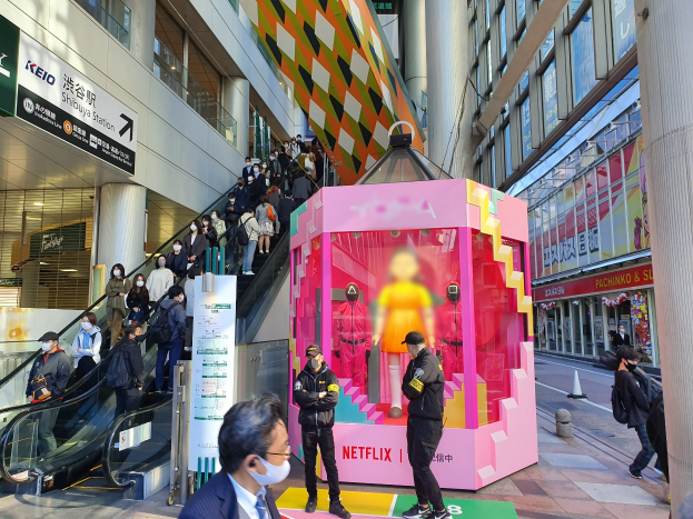 Eine Gruppe von Menschen vor einem Gebäude mit Glaswänden, Texttafeln, einige tragen Masken, in der Nähe einer Rolltreppe; eine pinkfarbene Box in der Mitte zeigt "Netflix's new Netflix-Booth in Tokyo" mit Schaufensterpuppen darin.