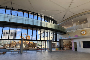 Innenansicht eines großen Gebäudes mit zahlreichen Fenstern, Deckenbeleuchtung, einer Wanduhr, einem Fernseher, einem Bildschirm, Holzkonstruktionen, einem Holzzaun, umgeben von Gebäuden, Bäumen und einem bewölkten Himmel, als Haupteingang zu einem neuen Kinderspielplatz.