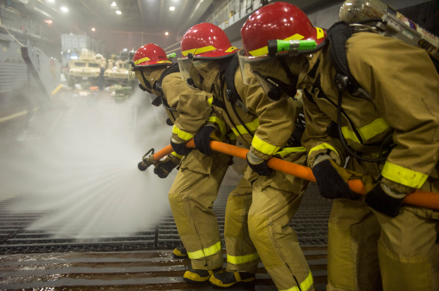Feuerwehrleute in Helmen und Handschuhen mit Rohren, die einen Löschwagen mit Wasser bespritzen, mit Lampen, Geländern und Tafeln mit Text im Hintergrund.