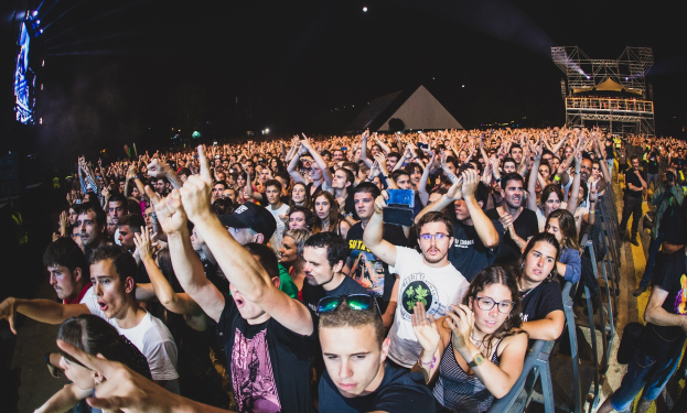 Menschenmenge auf einem Musikfestival mit erhobenen Händen, sitzend und stehend, nahe einem Metallzaun, Gebäuden und Lichtern unter einem dunklen Himmel.