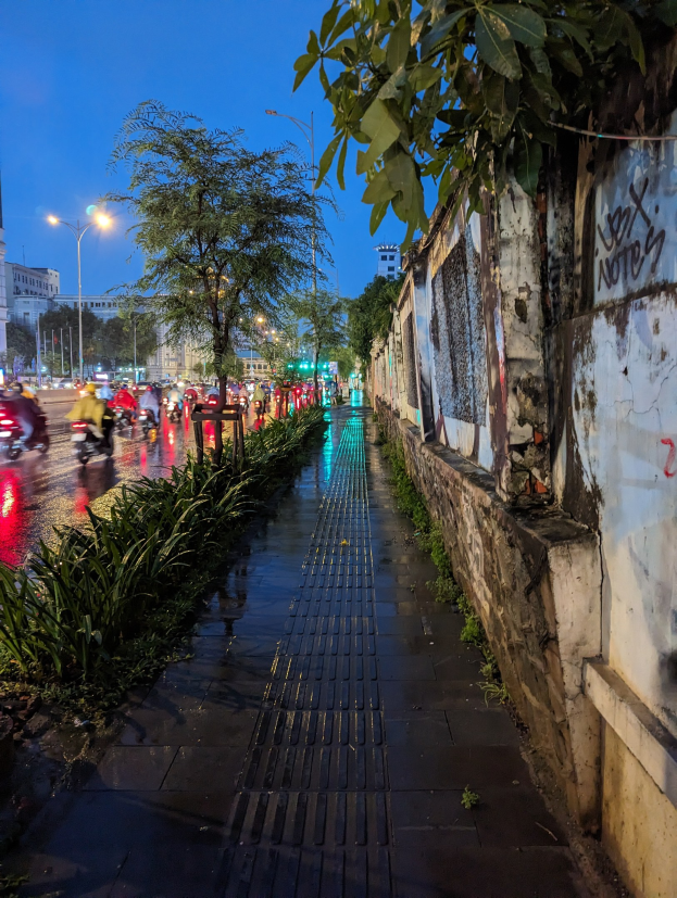 Stadtstraße bei Nacht mit Graffiti an der Seite, beleuchtet von Straßenlaternen, fahrende Kraftfahrzeuge, Bäume und Pflanzen an den Seiten, Gebäude und ein sternenklarer Himmel im Hintergrund.