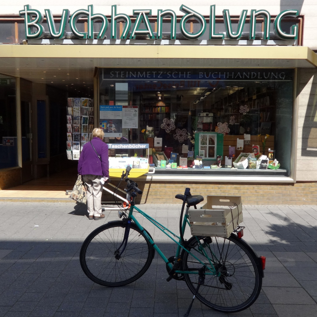 Eine Frau steht neben einem Fahrrad, das vor einem Laden geparkt ist, mit einem Fahrradrad im Vordergrund und mit Büchern und anderen Gegenständen durch die Gläserscheibe des Ladens sichtbar.